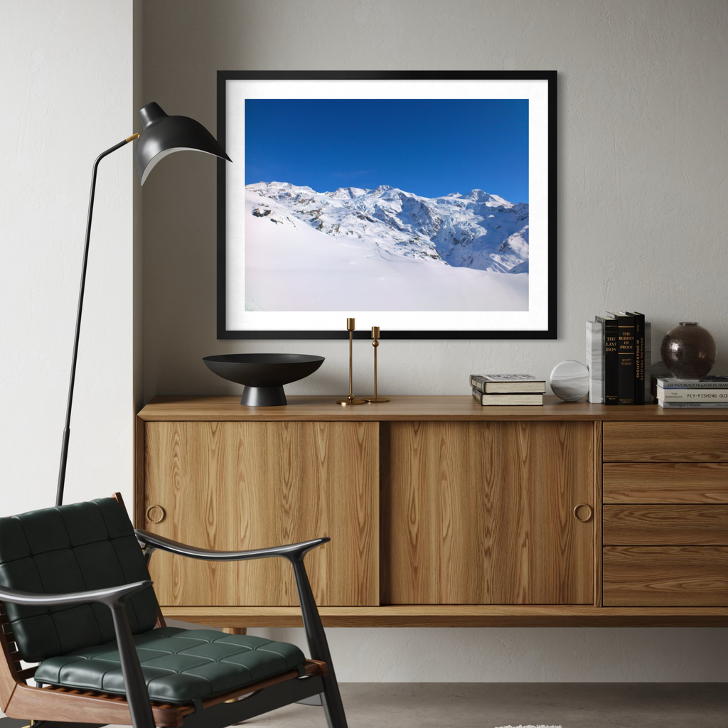 A framed art print of a snowy mountain landscape under a clear blue sky, displayed on a light gray wall above a wooden sideboard in a modern living room setting.