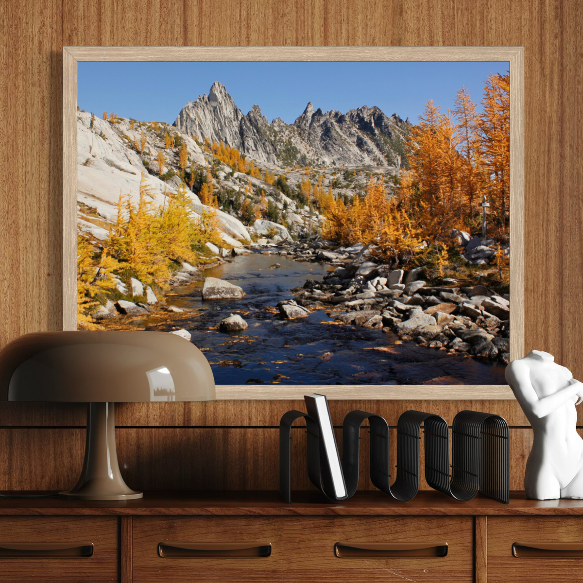 A wooden framed landscape art print depicting a rocky mountain valley with a stream and golden autumn trees, displayed on a wooden wall above a console table with a lamp and decorative objects.