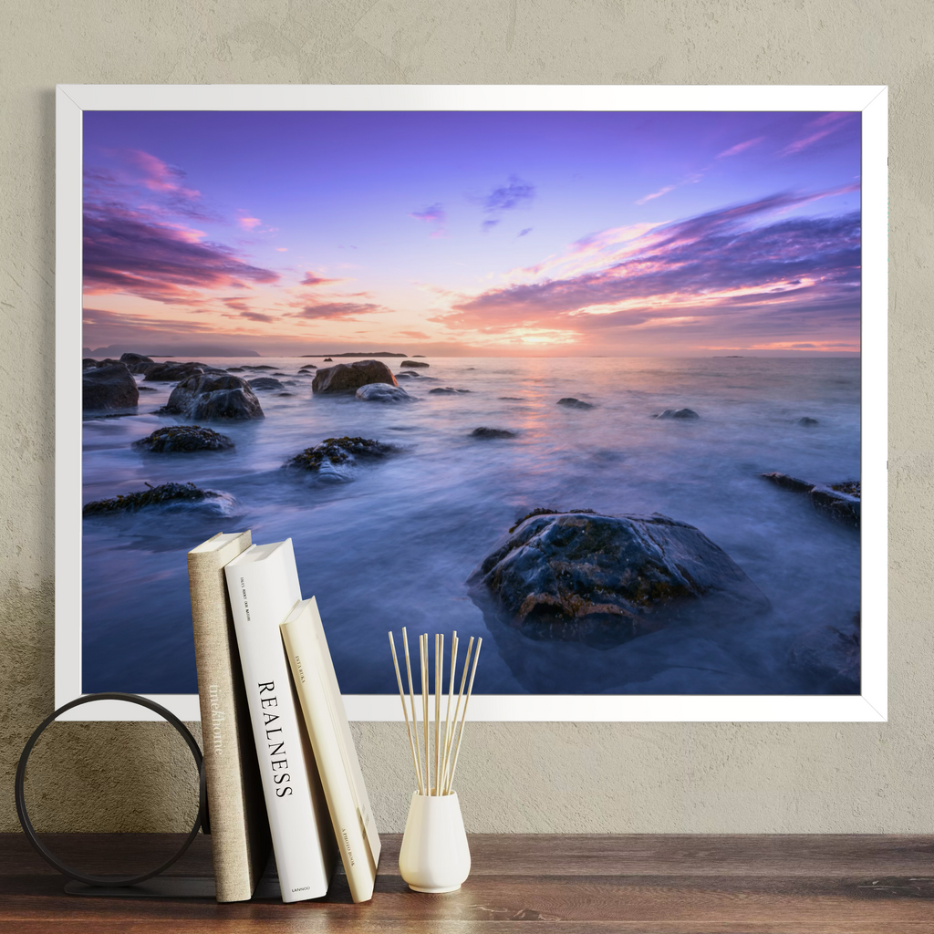 A framed wall art print showing a rocky ocean sunset in purple, pink, blue, and orange tones, displayed above a wooden surface with books and a reed diffuser against a textured beige wall.