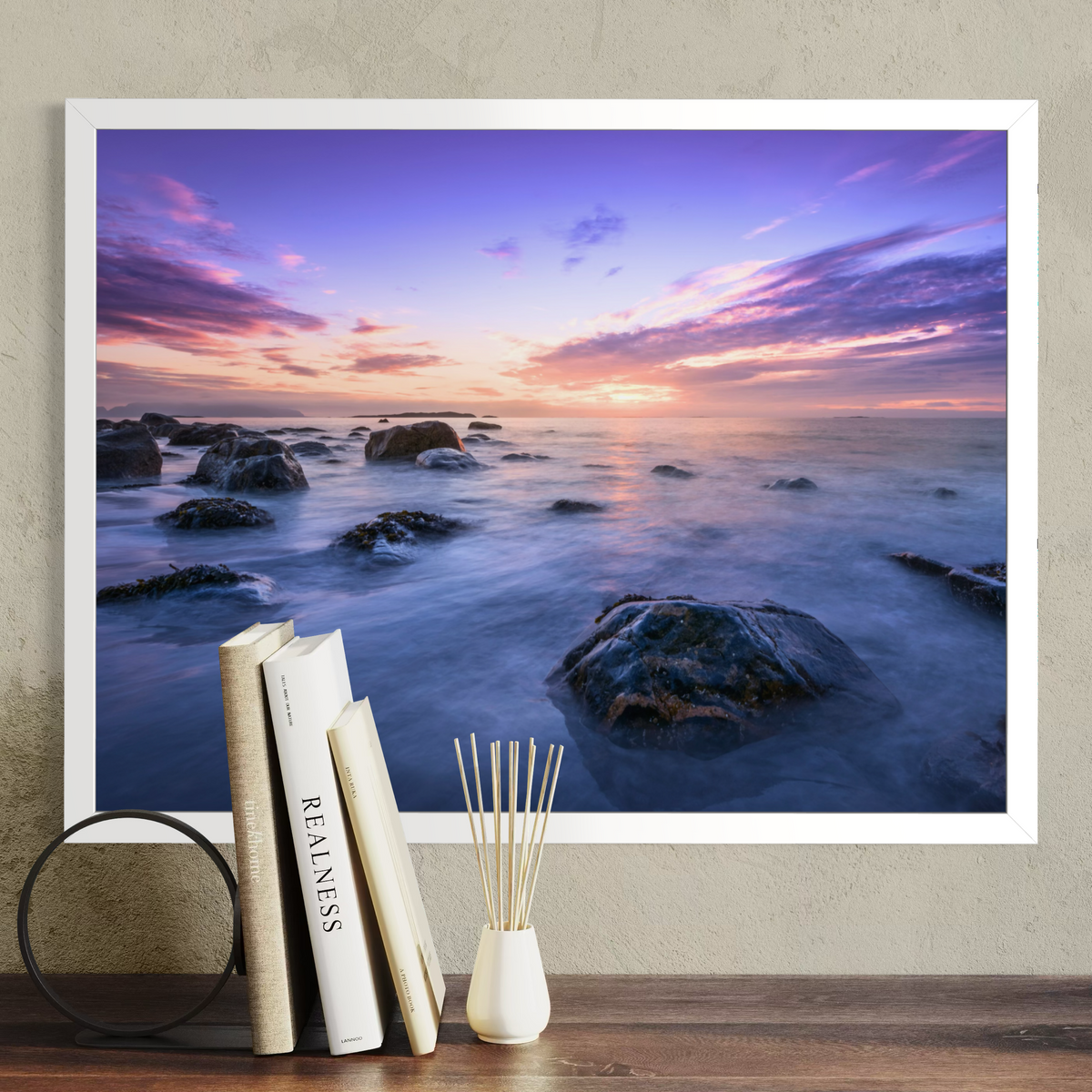 A framed wall art print showing a rocky ocean sunset in purple, pink, blue, and orange tones, displayed above a wooden surface with books and a reed diffuser against a textured beige wall.
