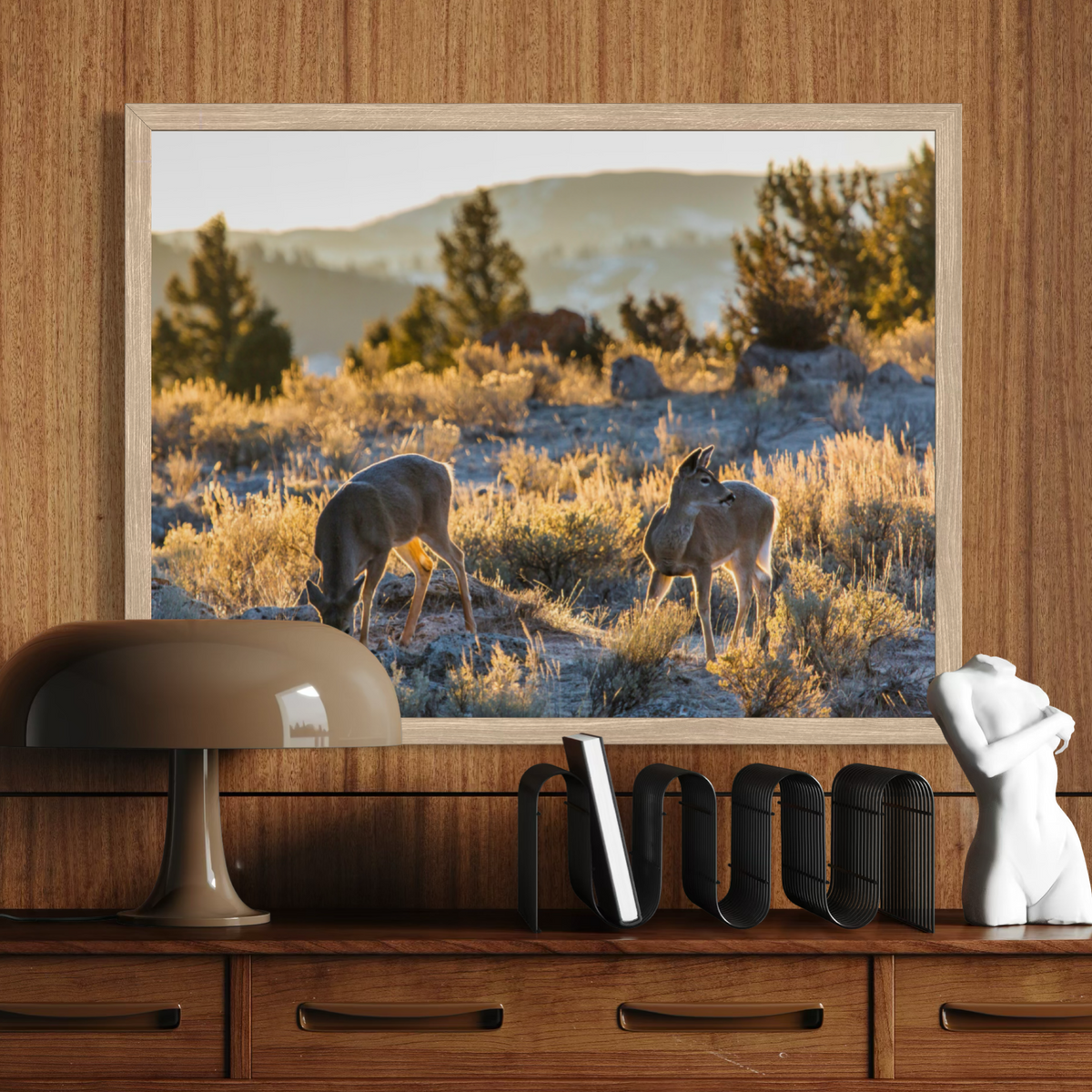 A wooden framed art print depicting two deer grazing in a sunlit desert landscape, displayed on a wooden console table against a matching wood-paneled wall, styled with a lamp, books, and a white bust.