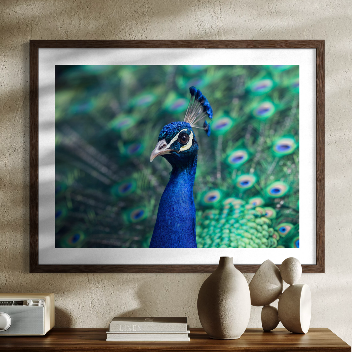 A framed wall art print featuring a close-up photograph of a peacock with vivid blue neck and green peacock feathers in the background, displayed above a console table with decorative vases and books.
