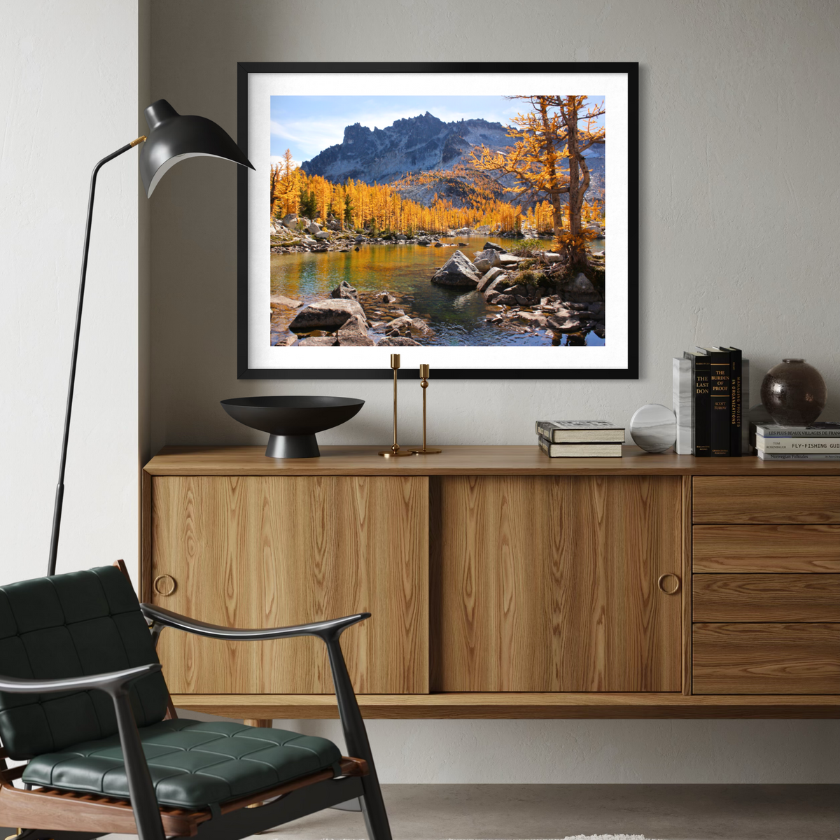 A black framed landscape art print of a mountain river scene with golden autumn trees is displayed on a light gray wall above a wooden credenza in a modern living room setting.