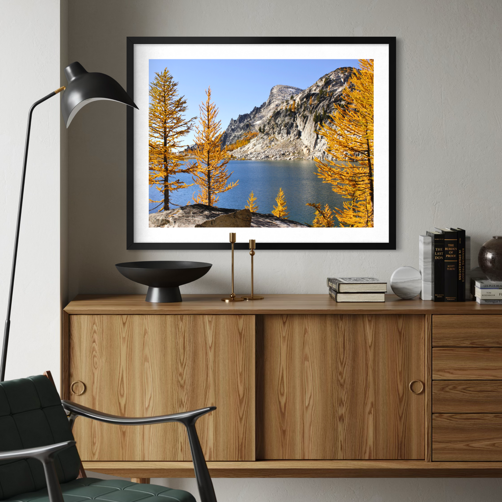A black framed landscape art print of a mountain lake with golden larch trees, displayed on a gray wall above a mid-century modern wooden credenza in a styled room.