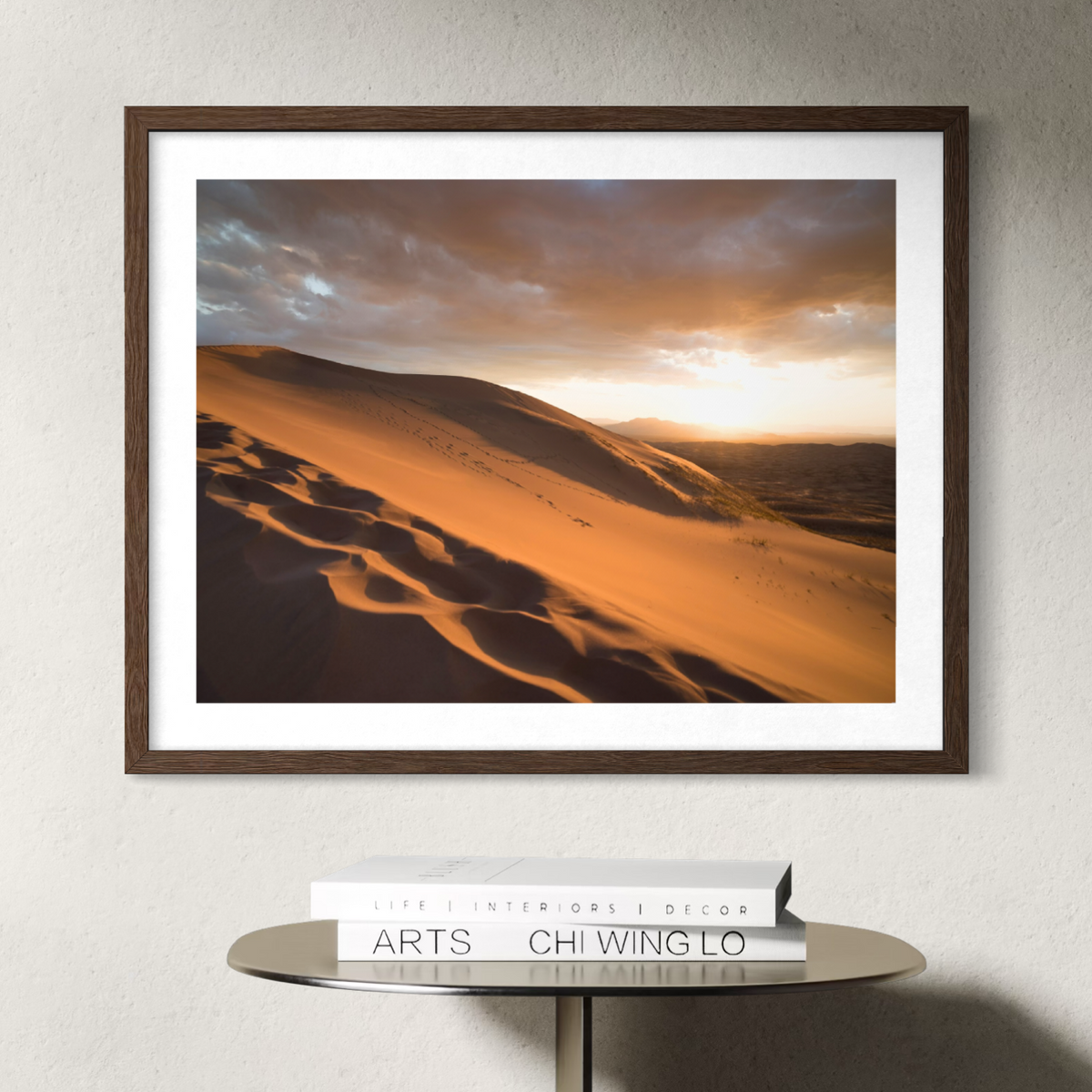 A framed landscape art print depicting golden desert sand dunes under a dramatic sunset sky, styled on a round table above a stack of books against a textured wall.
