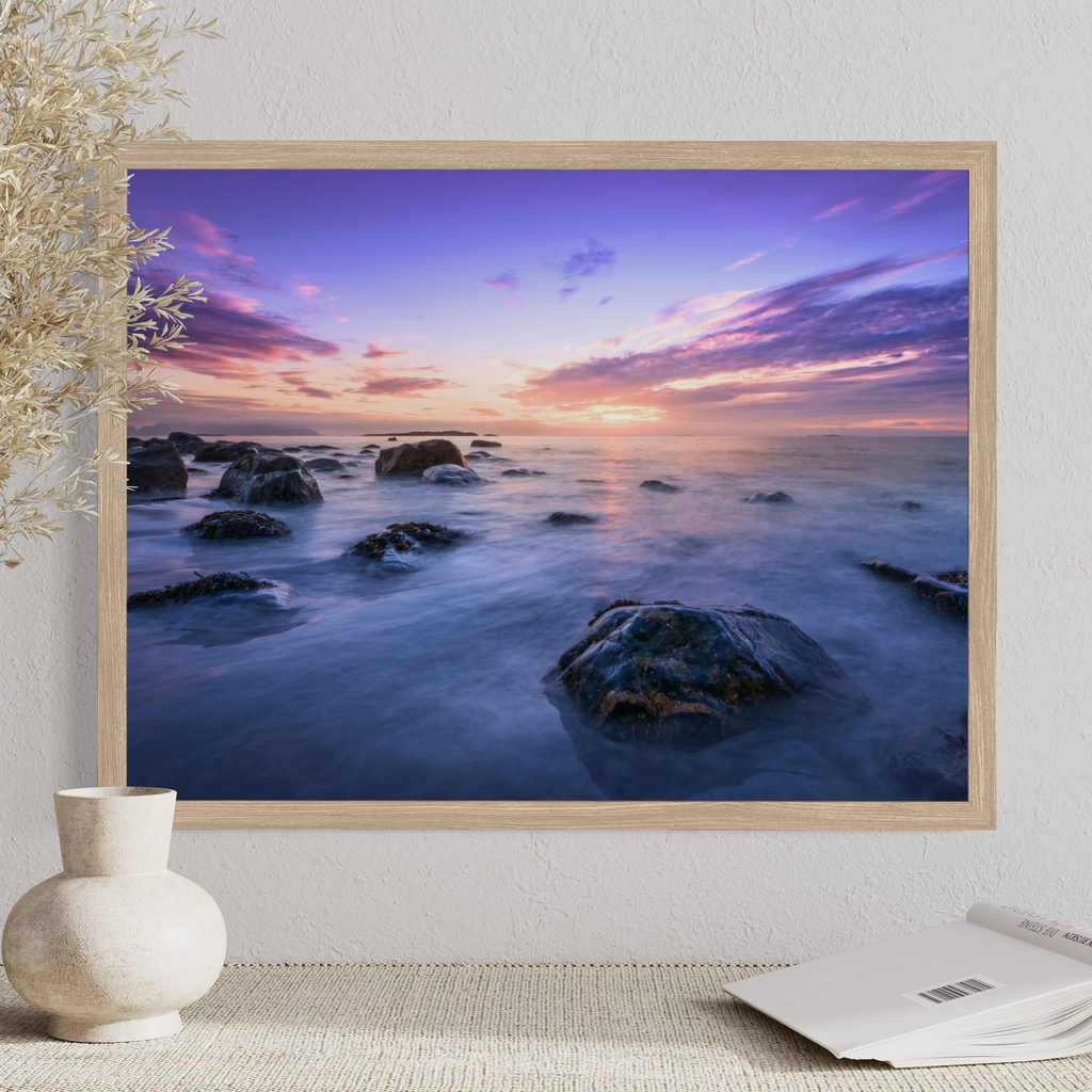 A framed landscape art print of a rocky ocean sunset, displayed on a textured wall above a table with a vase and an open book.
