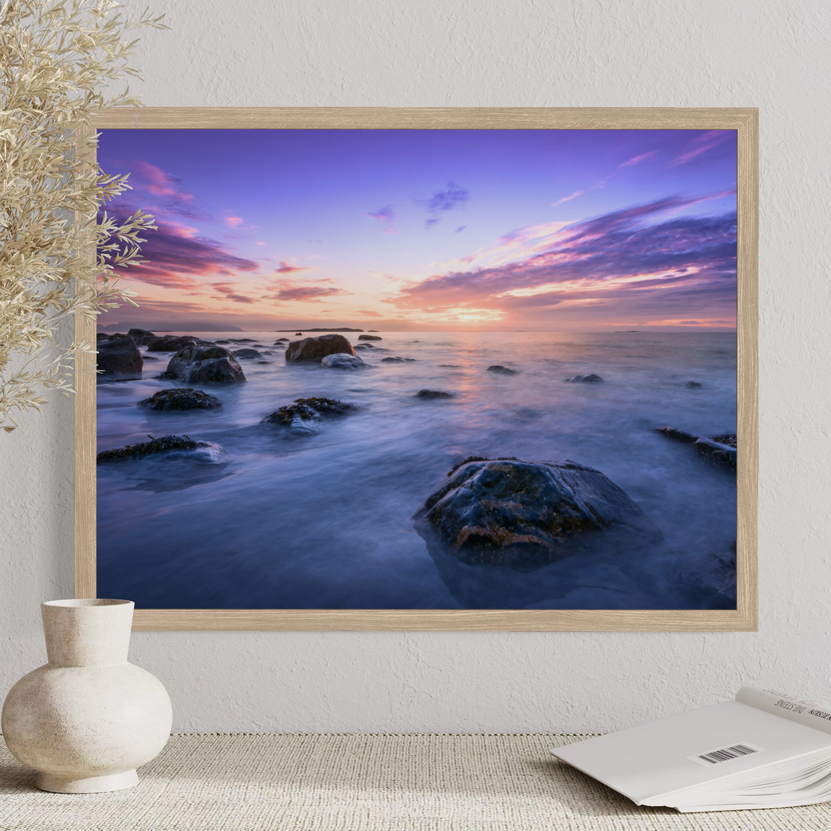A framed landscape art print of a rocky ocean sunset, displayed on a textured wall above a table with a vase and an open book.