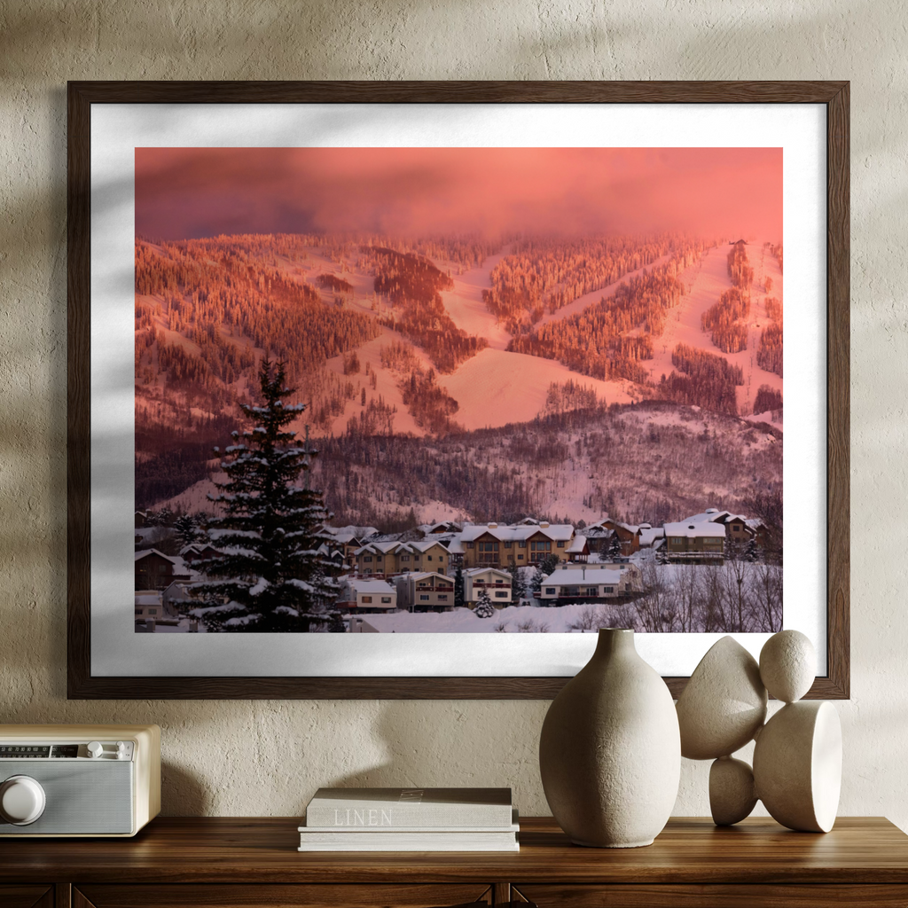 A framed wall art print depicting a snowy mountain village under a warm pink-orange sky, displayed above a wooden console with decorative objects against a textured beige wall.
