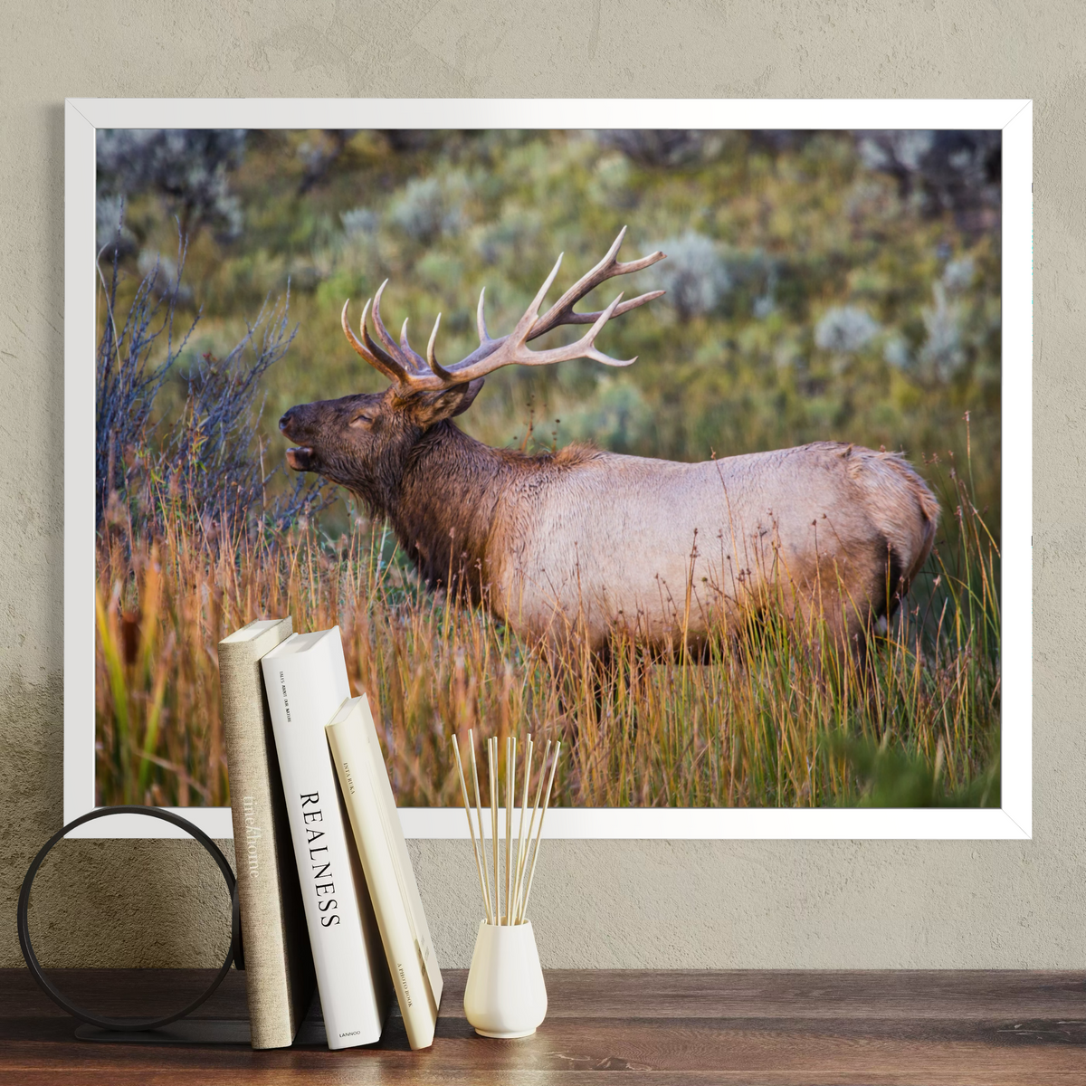 A white framed wall art print showing a large elk standing in tall grass, displayed on a beige wall above a wooden surface with books and a reed diffuser in front.