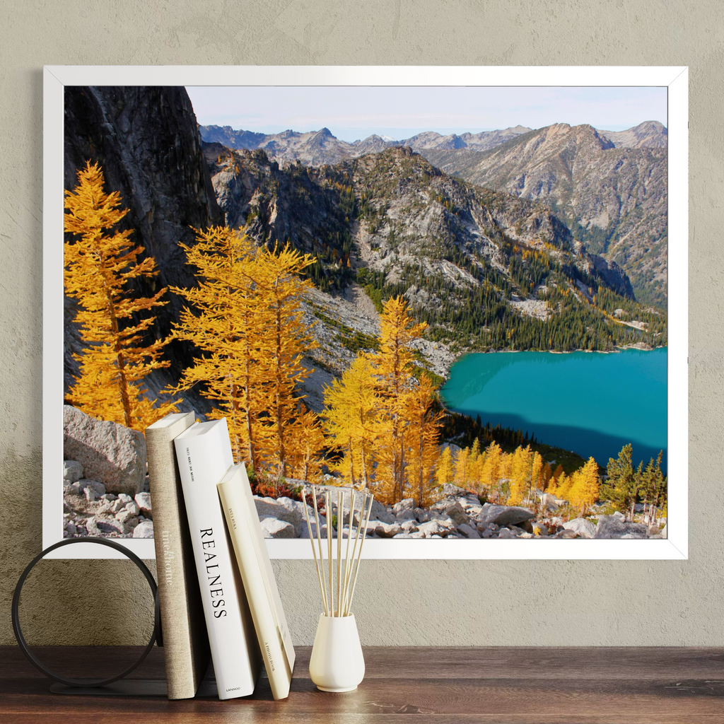 A white framed landscape art print depicting a mountain range with golden yellow larchwood trees and a turquoise lake, displayed on a wooden shelf next to books and a small vase.