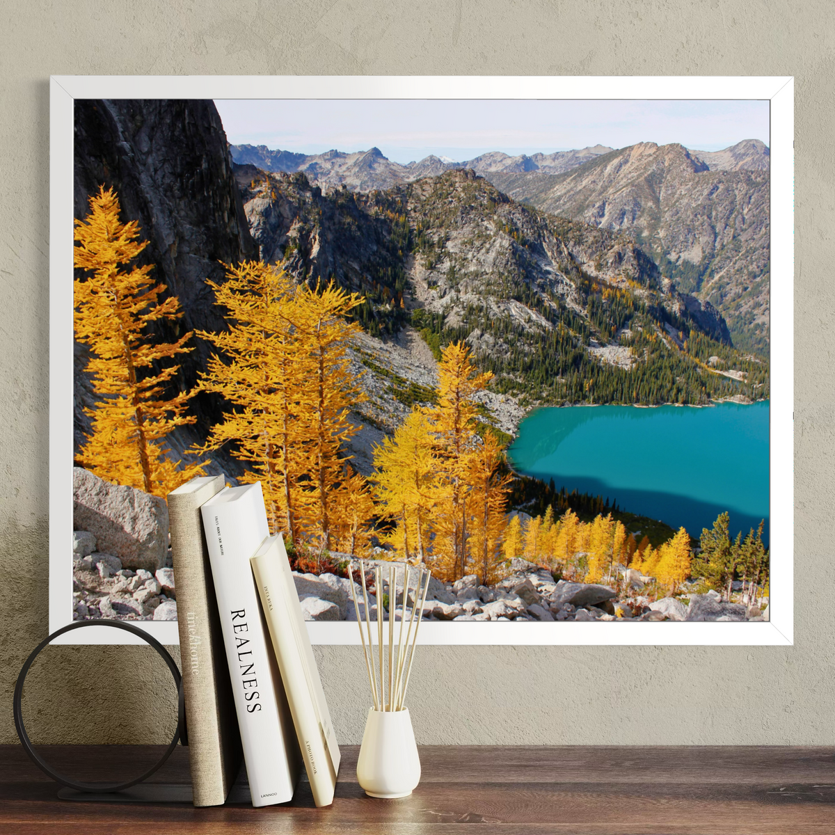 A white framed landscape art print depicting a mountain range with golden yellow larchwood trees and a turquoise lake, displayed on a wooden shelf next to books and a small vase.