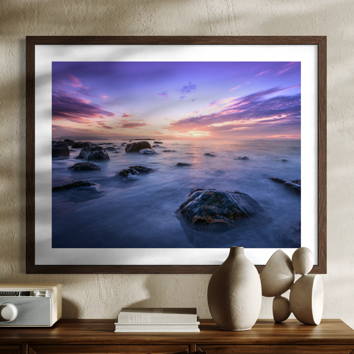 A wooden framed wall art print showing a coastal seascape at sunset with purple, pink, and blue sky, rocks, and misty water, displayed above a wooden console with decorative objects.