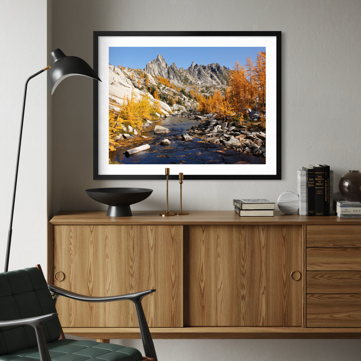 A black wooden framed landscape art print of a mountain river scene with autumn foliage, displayed on a gray wall above a wooden console table in a modern living room setting.
