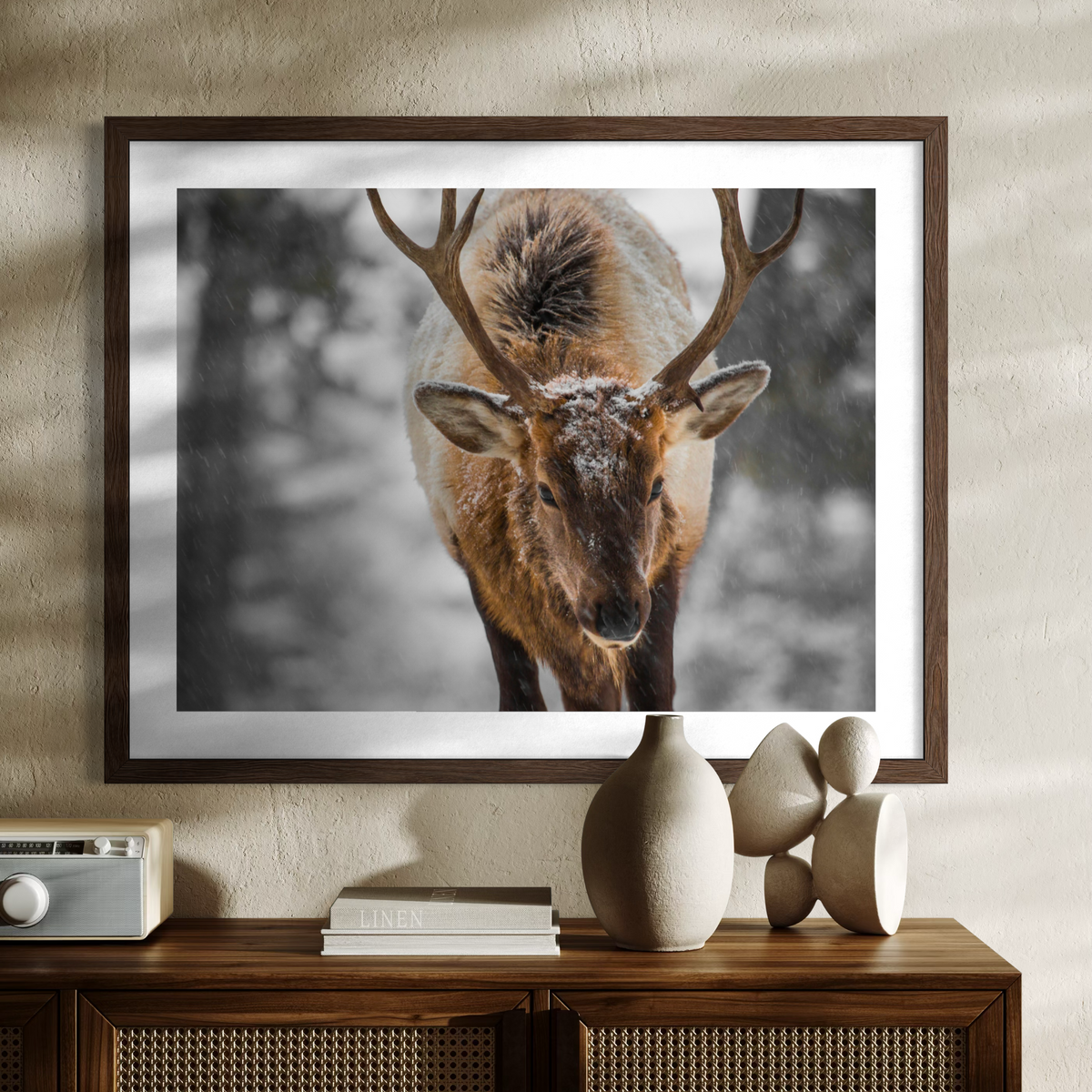 A wooden framed wall art print showing a close-up photograph of a deer with large antlers in a snowy outdoor scene, displayed above a wooden console table with decorative objects.