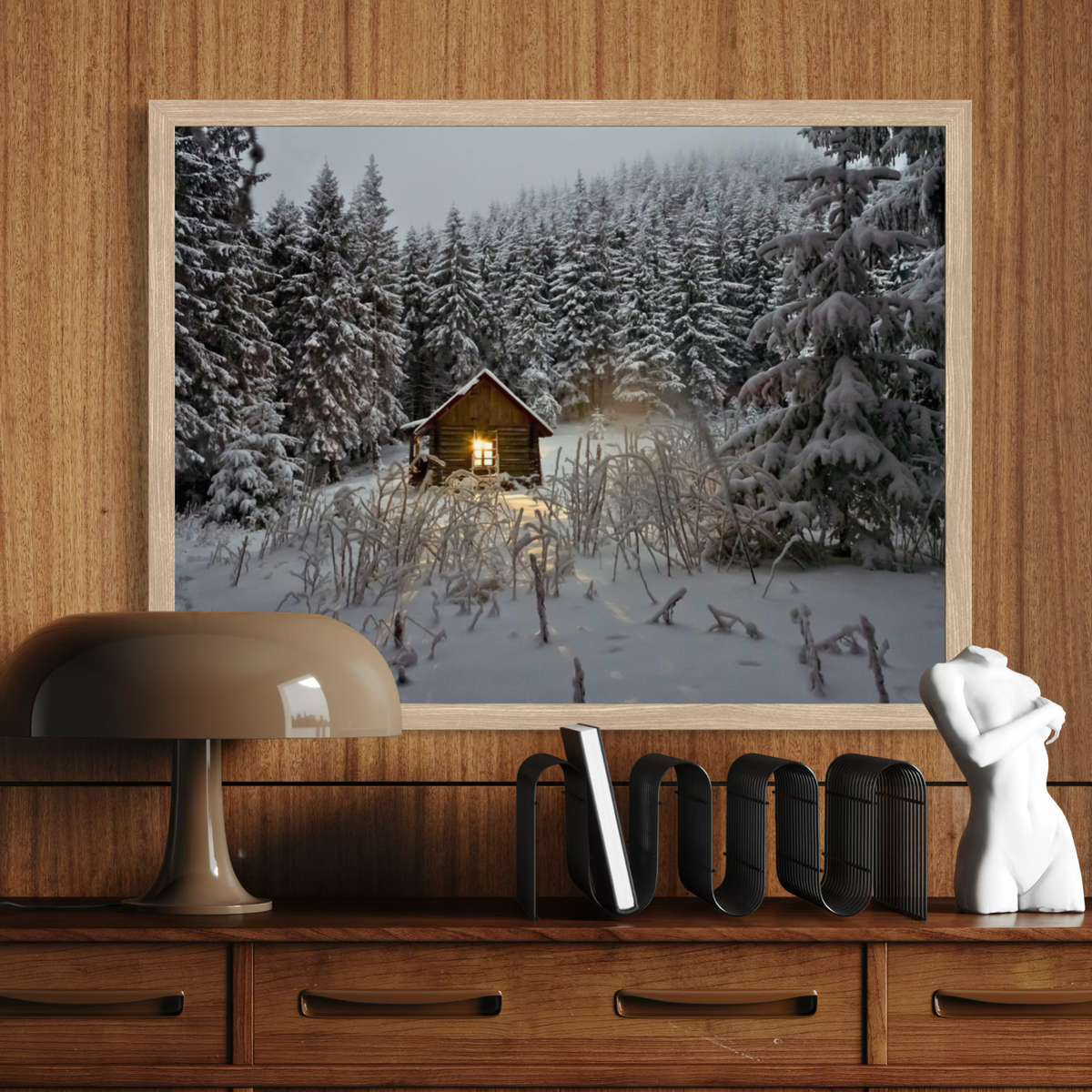 A framed winter forest print featuring a small cabin with a glowing window in a snow-covered woodland, displayed on a wooden console above furniture.