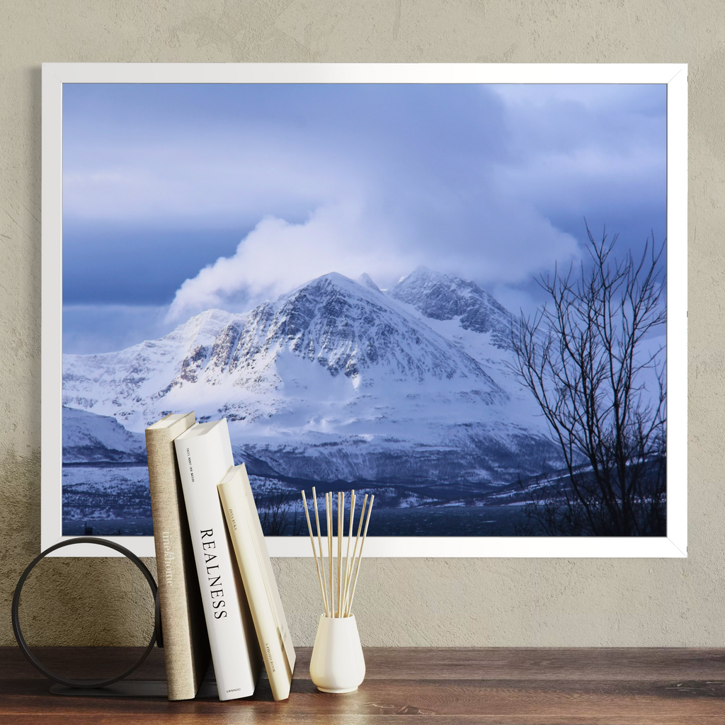 A large framed wall art print showing a snowy mountain landscape under a blue cloudy sky, displayed on a beige wall above a wooden surface with books, a small vase, and a diffuser.