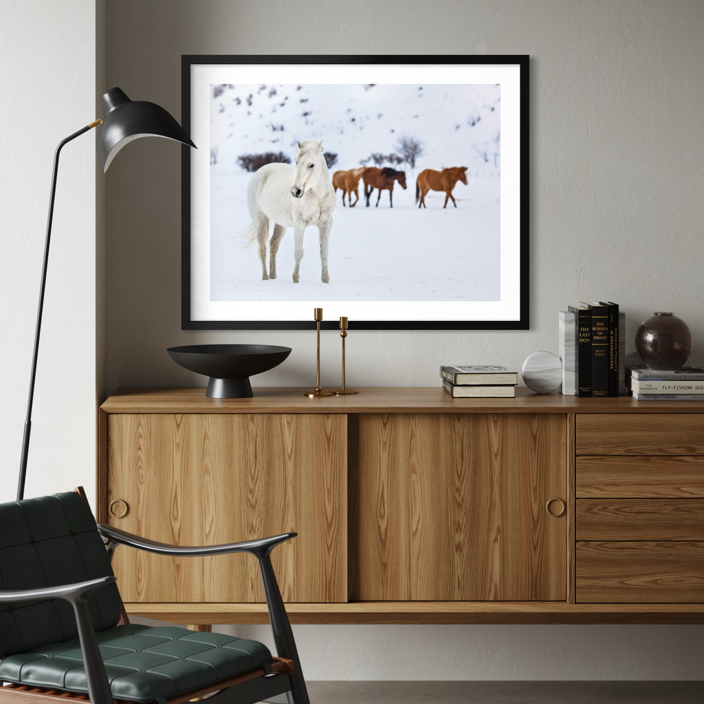 A framed art print of horses in a snowy landscape, displayed on a wooden cabinet in a modern living room setting.