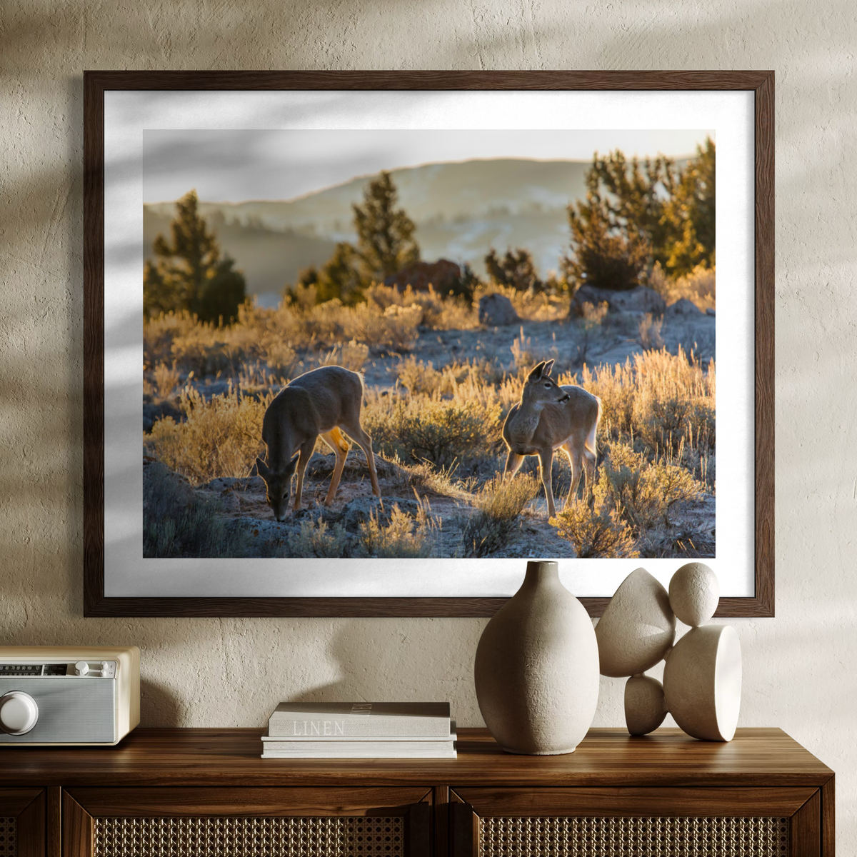 A wooden framed art print depicting two antelope grazing in a golden grassy landscape, displayed on a wooden console table with decorative ceramic vases and books against a textured beige wall.