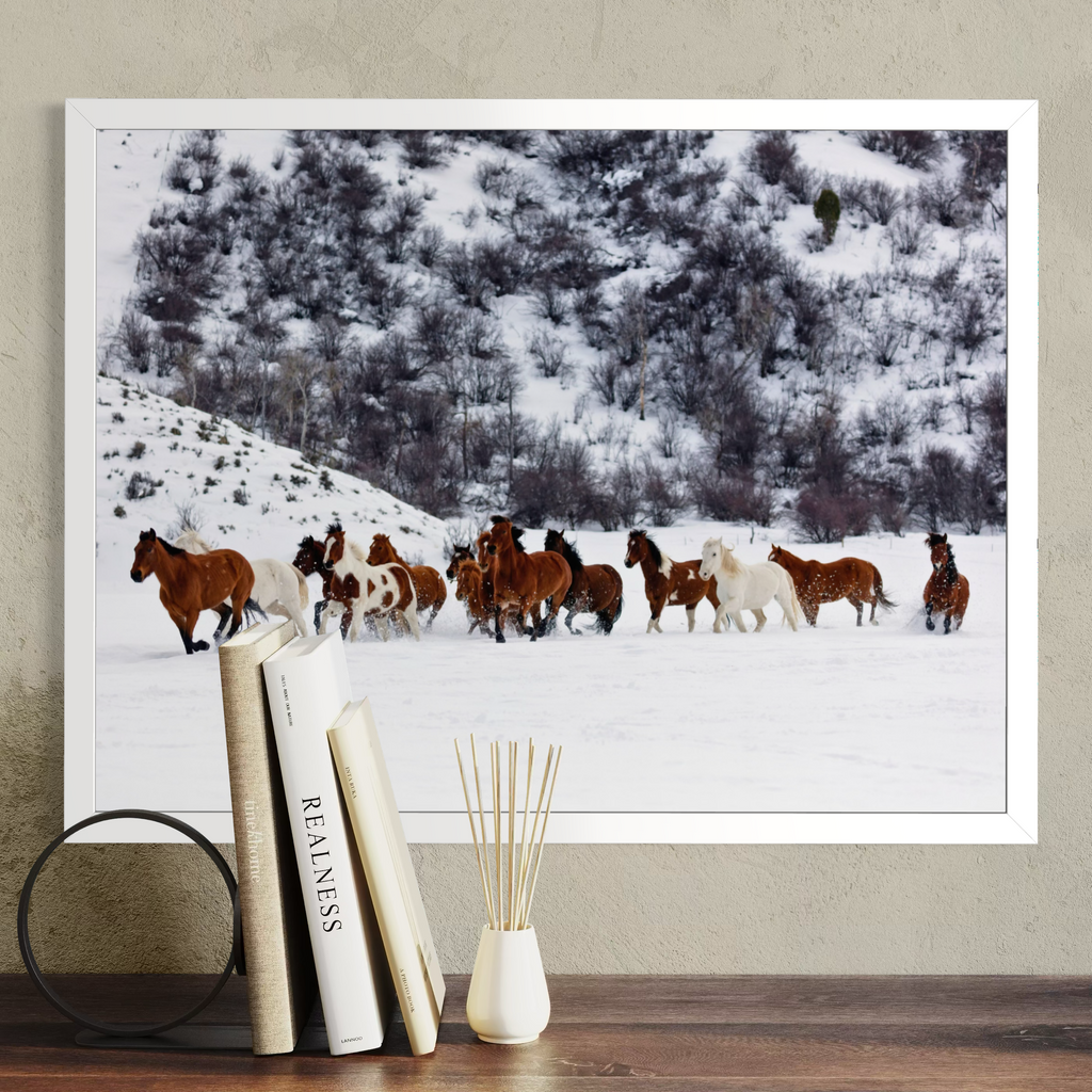 A framed wall art print showing a herd of horses running through deep snow, displayed above a wooden table with books and a reed diffuser. The print is in a white frame against a textured beige wall.