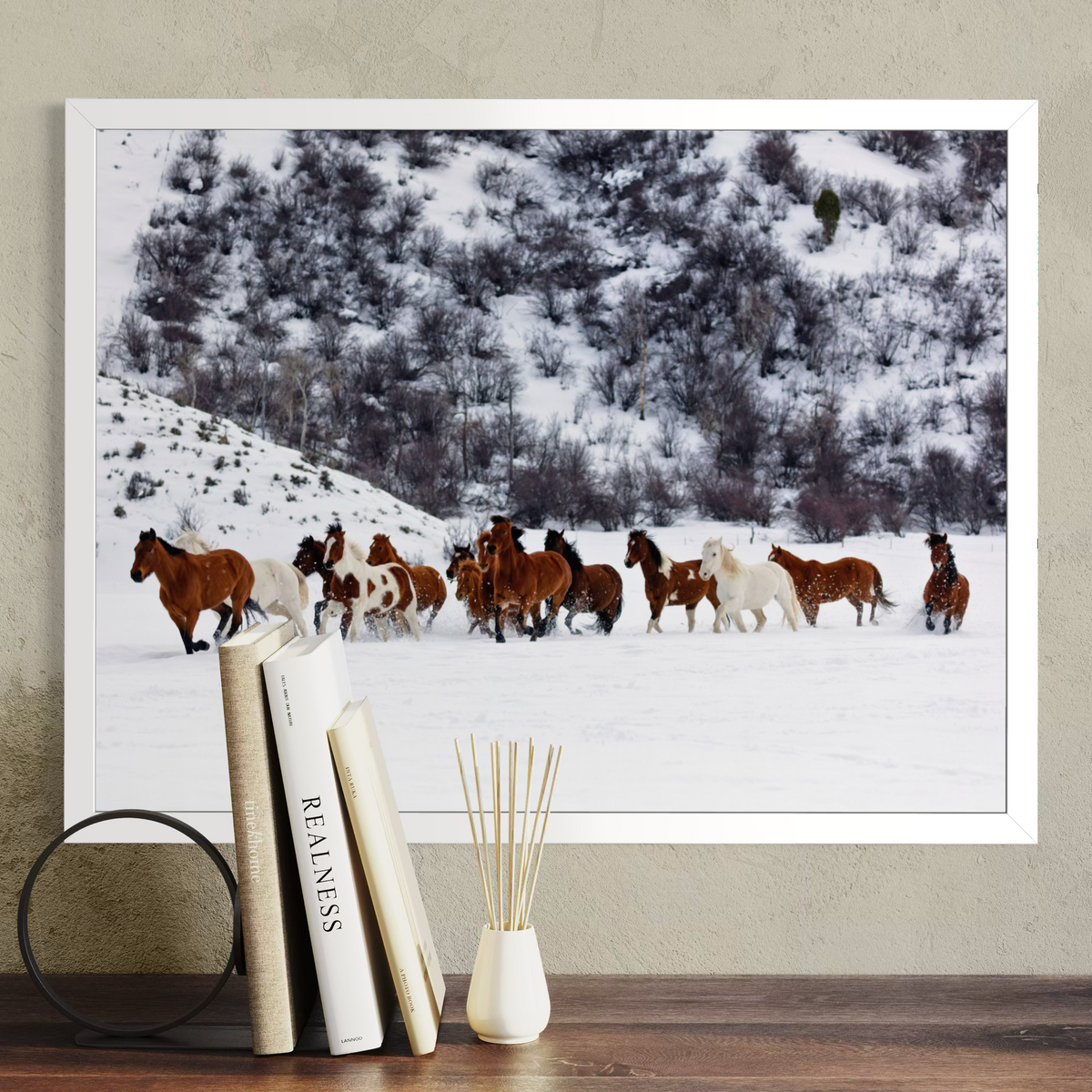 A framed wall art print showing a herd of horses running through deep snow, displayed above a wooden table with books and a reed diffuser. The print is in a white frame against a textured beige wall.