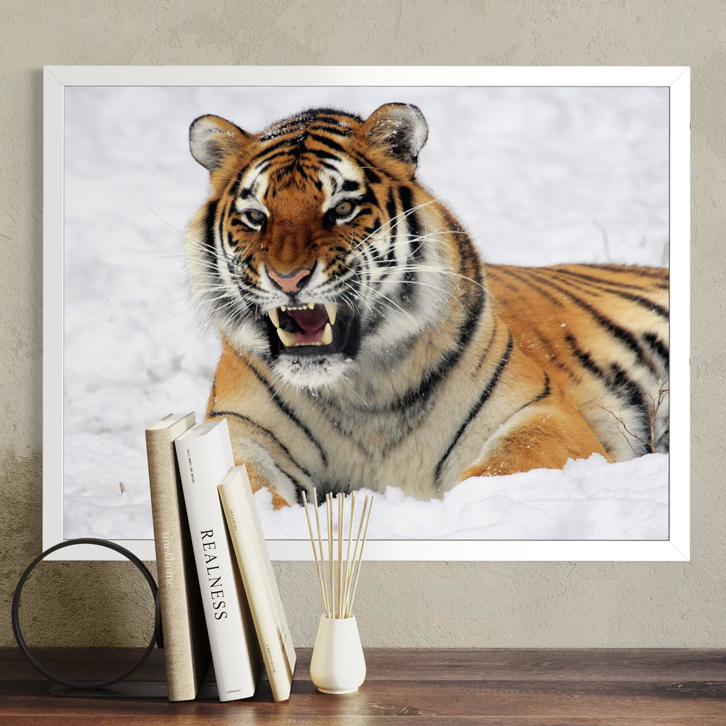 A white framed wall art print showing a close-up tiger face and body in snow, displayed above a wooden surface with books and a small vase against a beige wall.