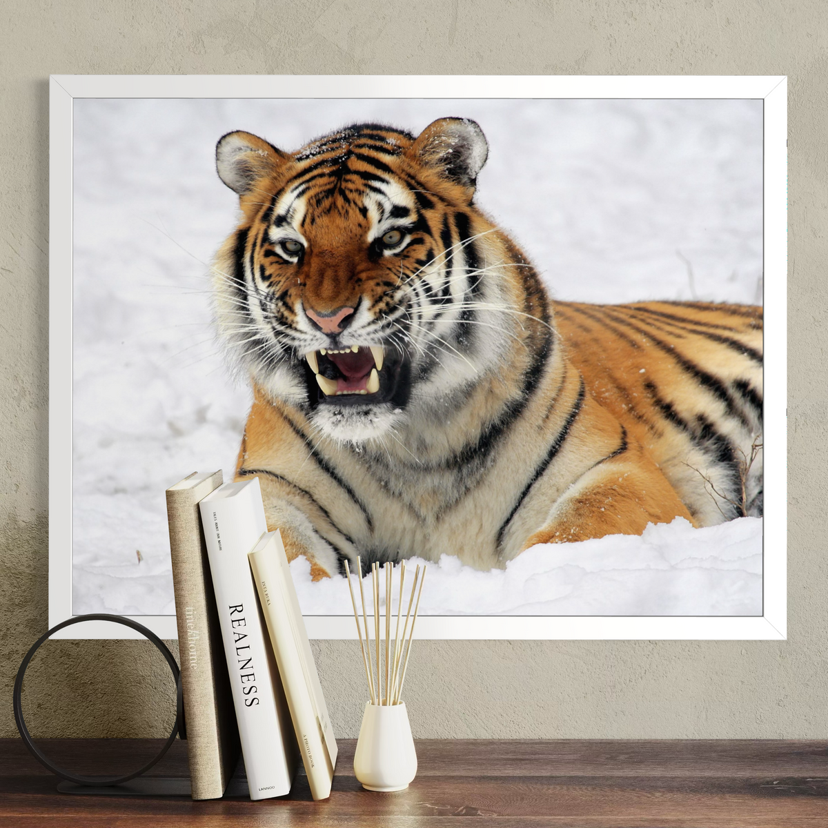 A white framed wall art print showing a close-up tiger face and body in snow, displayed above a wooden surface with books and a small vase against a beige wall.