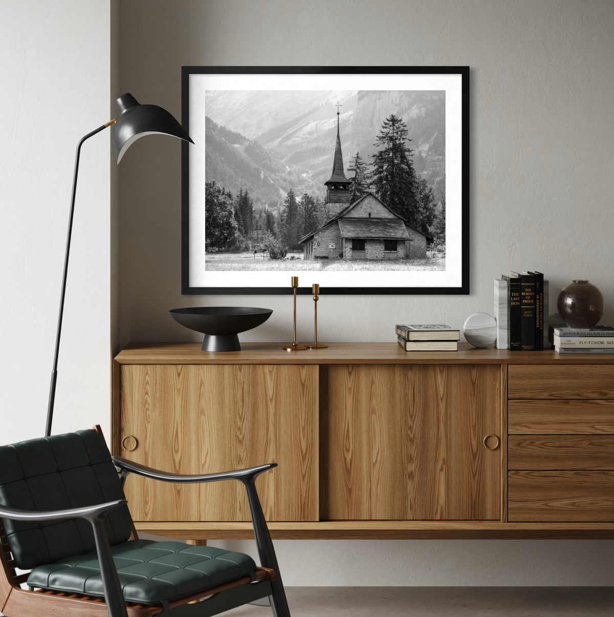 A black and white framed art print of a mountain church scene, displayed on a gray wall above a wooden credenza in a modern living room setting.