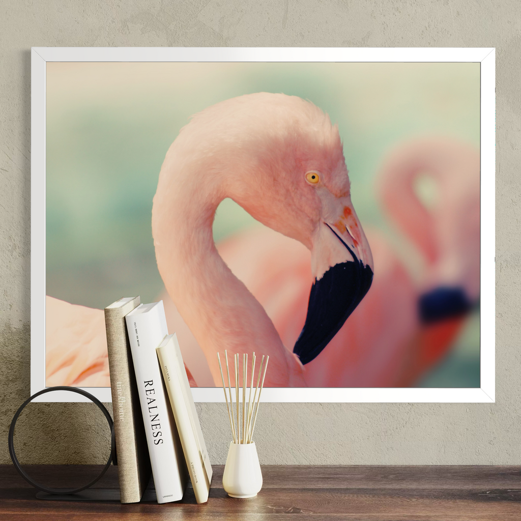 A framed art print of a pink flamingo hanging on a textured gray wall above a wooden table with books and a small vase.
