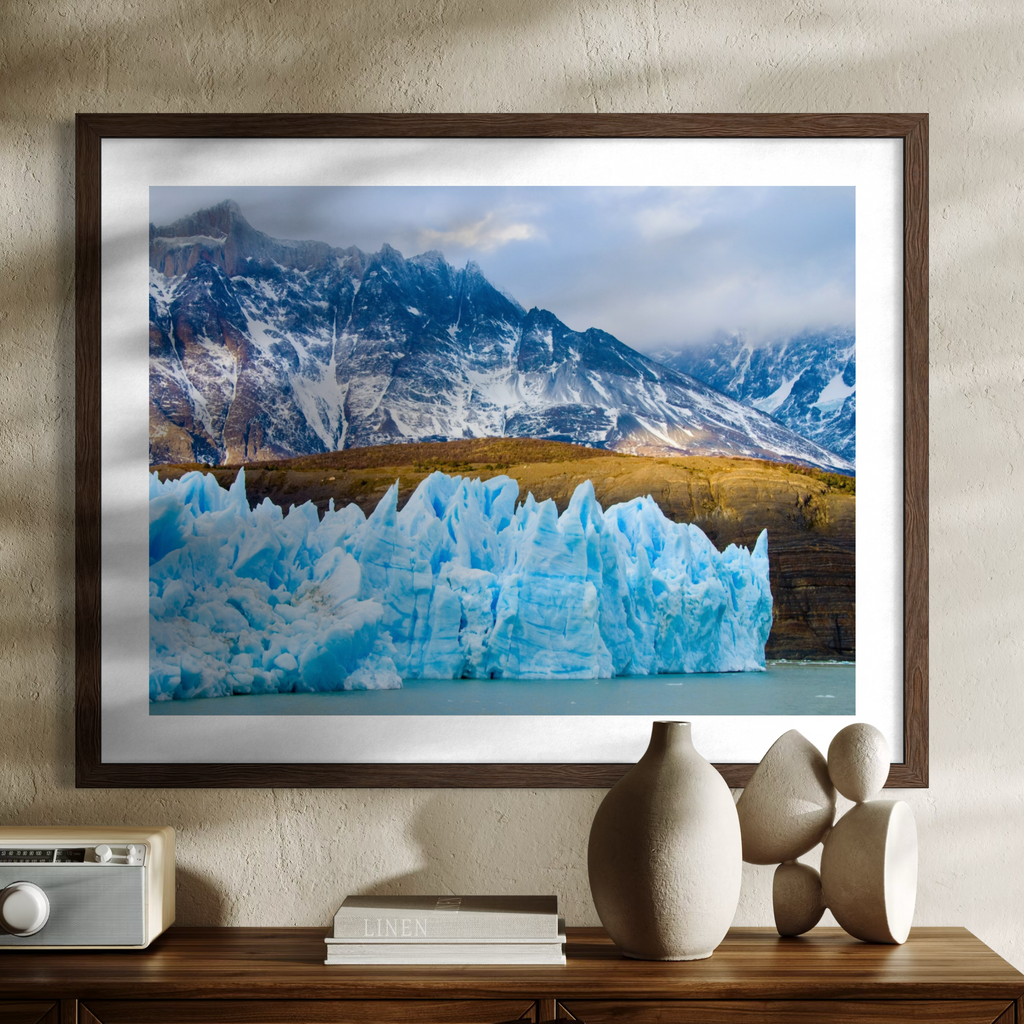 A framed landscape art print depicting a majestic blue glacier in a lake with snow-capped mountains in the background, styled on a wooden console table with decorative ceramic vases and a vintage radio against a textured beige wall.