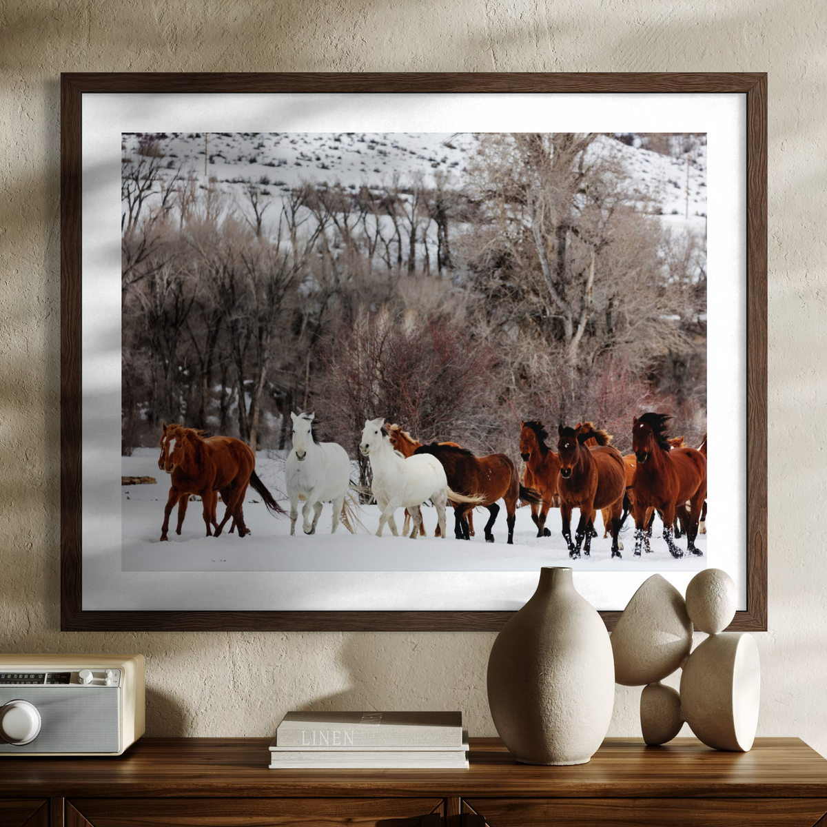 A framed wall art print showing a herd of horses in a snowy winter landscape, displayed above a wooden console table with decorative vases and books in a styled interior setting.