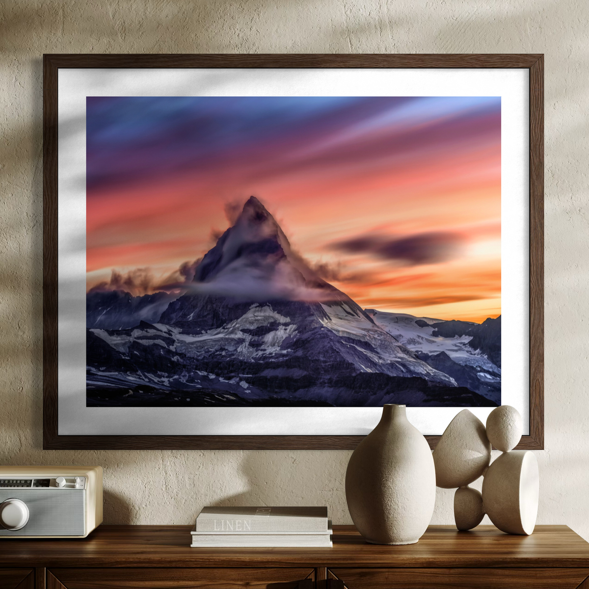 A framed art print of the Matterhorn mountain peak at sunset, displayed on a textured beige wall above a wooden console table with decorative ceramic vases and a radio.