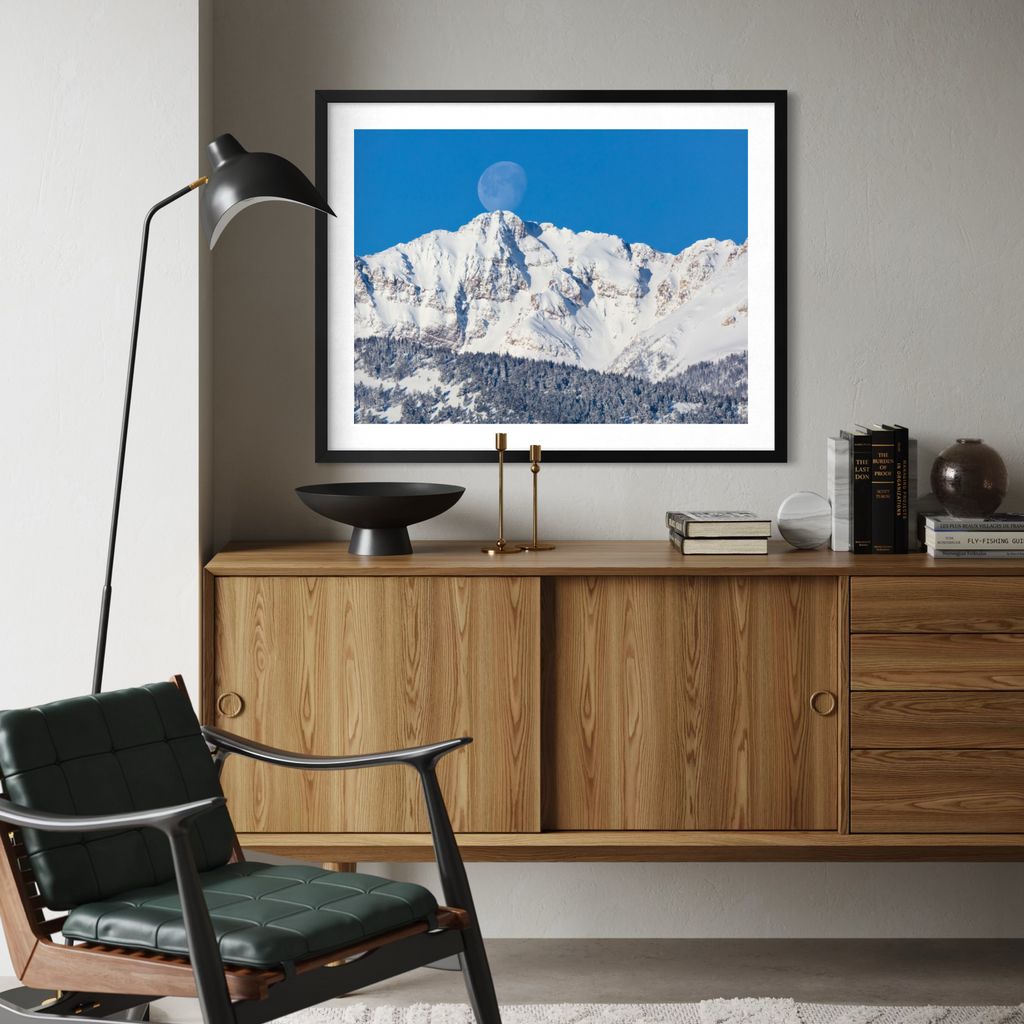 A framed art print of a snowy mountain landscape under a bright blue sky with a moon, displayed on a light gray wall above a mid-century modern wooden cabinet in a styled room.