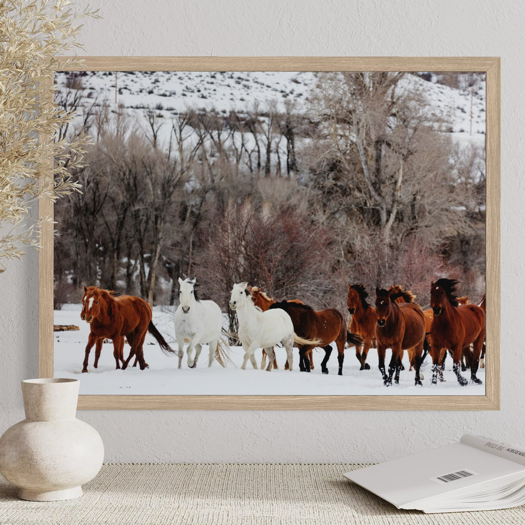 A framed art print depicting a herd of horses in a snowy landscape, displayed on a textured wall above a table with a vase and an open book.