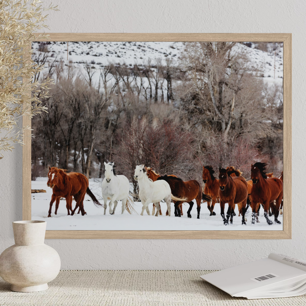 A framed art print depicting a herd of horses in a snowy landscape, displayed on a textured wall above a table with a vase and an open book.