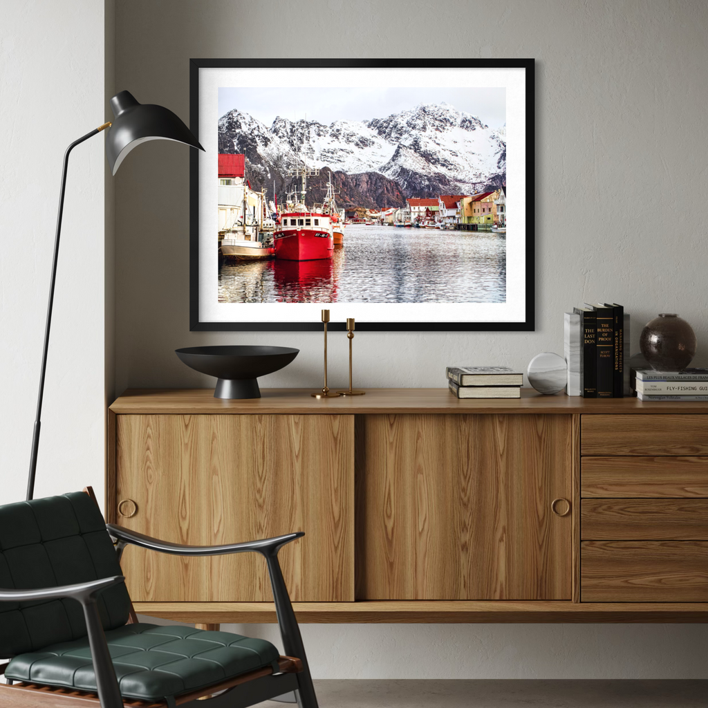 A framed art print depicting a scenic harbor with red boats and snow-capped mountains, displayed on a wooden cabinet in a modern living room setting.