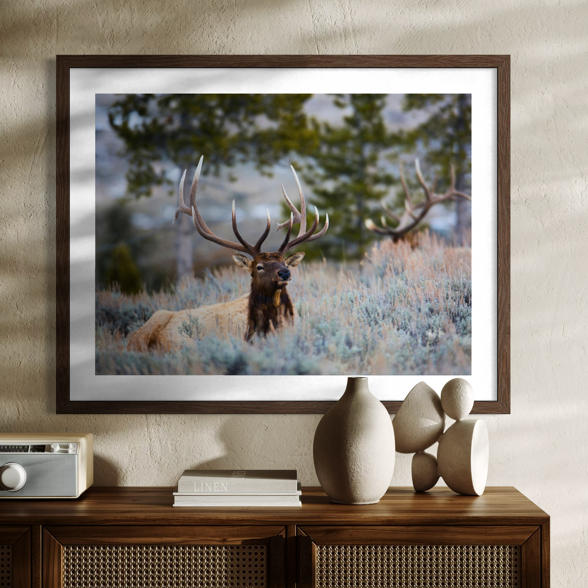 A wooden framed wall art print showing a large elk with antlers standing in a field of pale blue-gray grass, displayed above a wooden console table with decorative objects against a textured beige wall.