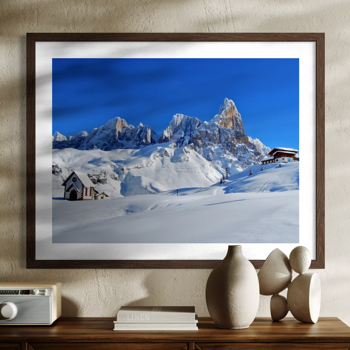 A framed art print depicting a snowy mountain landscape under a deep blue sky, styled on a beige wall above a wooden console table with decorative objects.