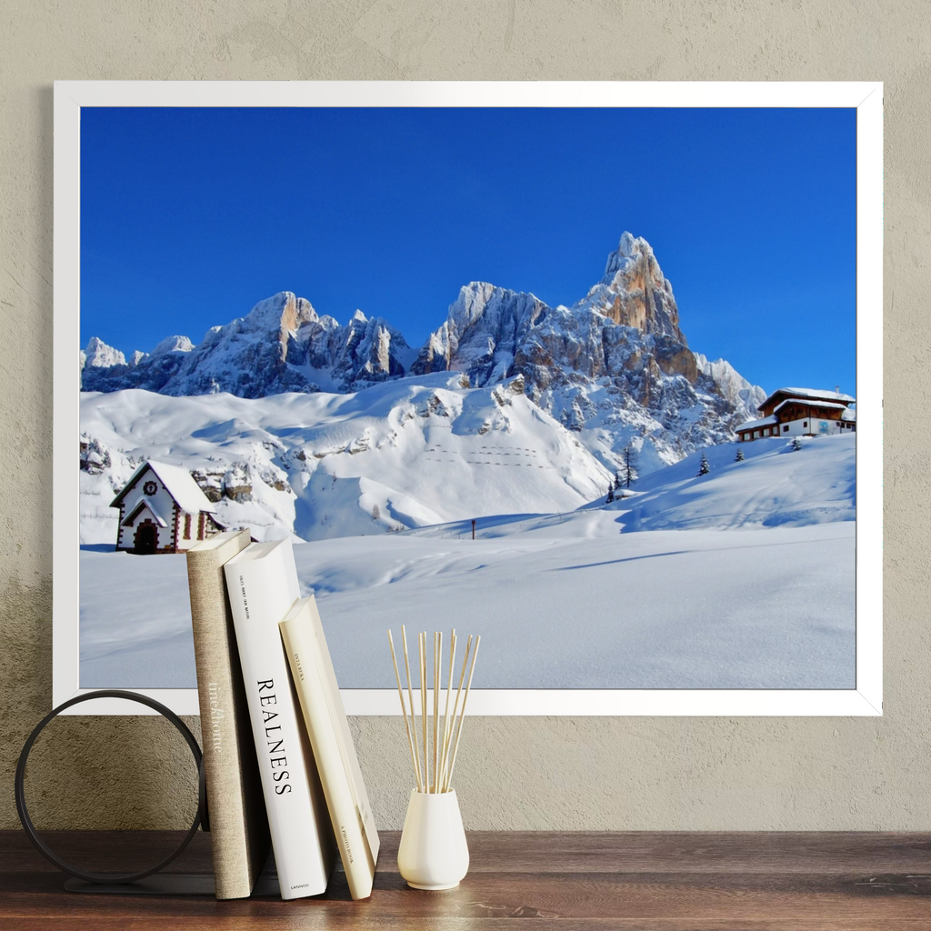 A framed wall art print showing a snowy mountain landscape under a vivid blue sky, displayed above a wooden surface with books and a small vase. The product being sold appears to be the framed print itself, not the decor props.