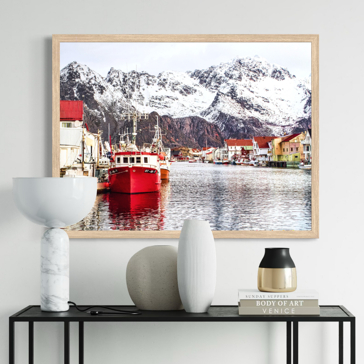 A framed art print depicting a scenic Norwegian fjord with a red fishing boat, colorful houses, and snow-capped mountains, displayed on a light gray wall above a console table with decorative vases and books.