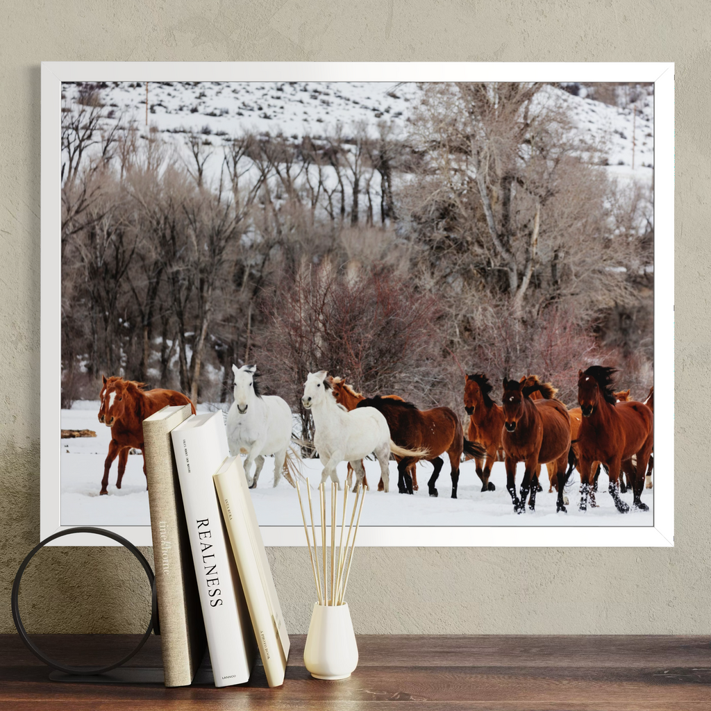A framed horizontal art print depicting a herd of horses running through a snowy landscape, styled on a wooden shelf next to books and a vase.