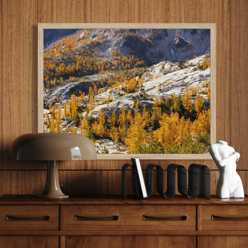 A wooden framed landscape art print depicting a mountain with golden-yellow larchwood trees and rocky slopes, displayed on a wooden console table in a styled room setting.