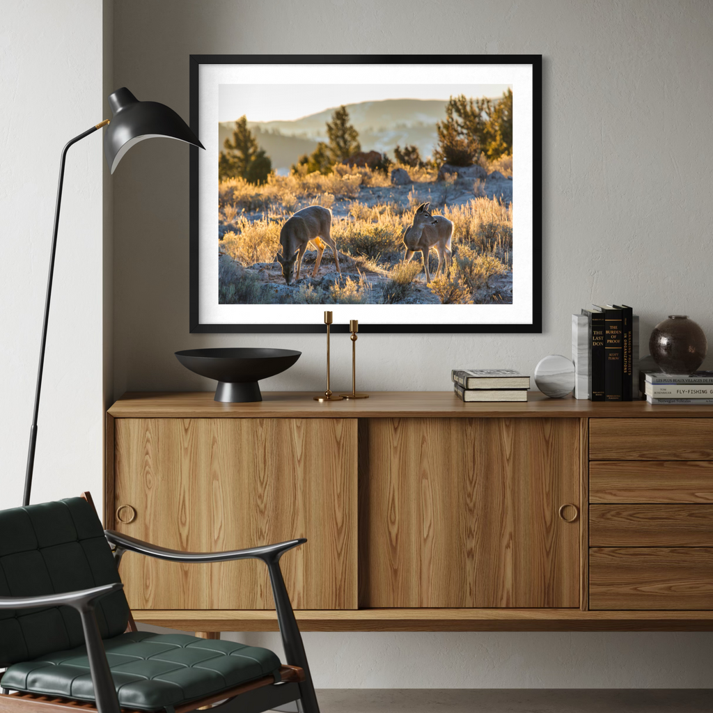 A framed landscape art print depicting two wild sheep in a golden grassy field at sunset, displayed on a wooden credenza in a modern living room setting.