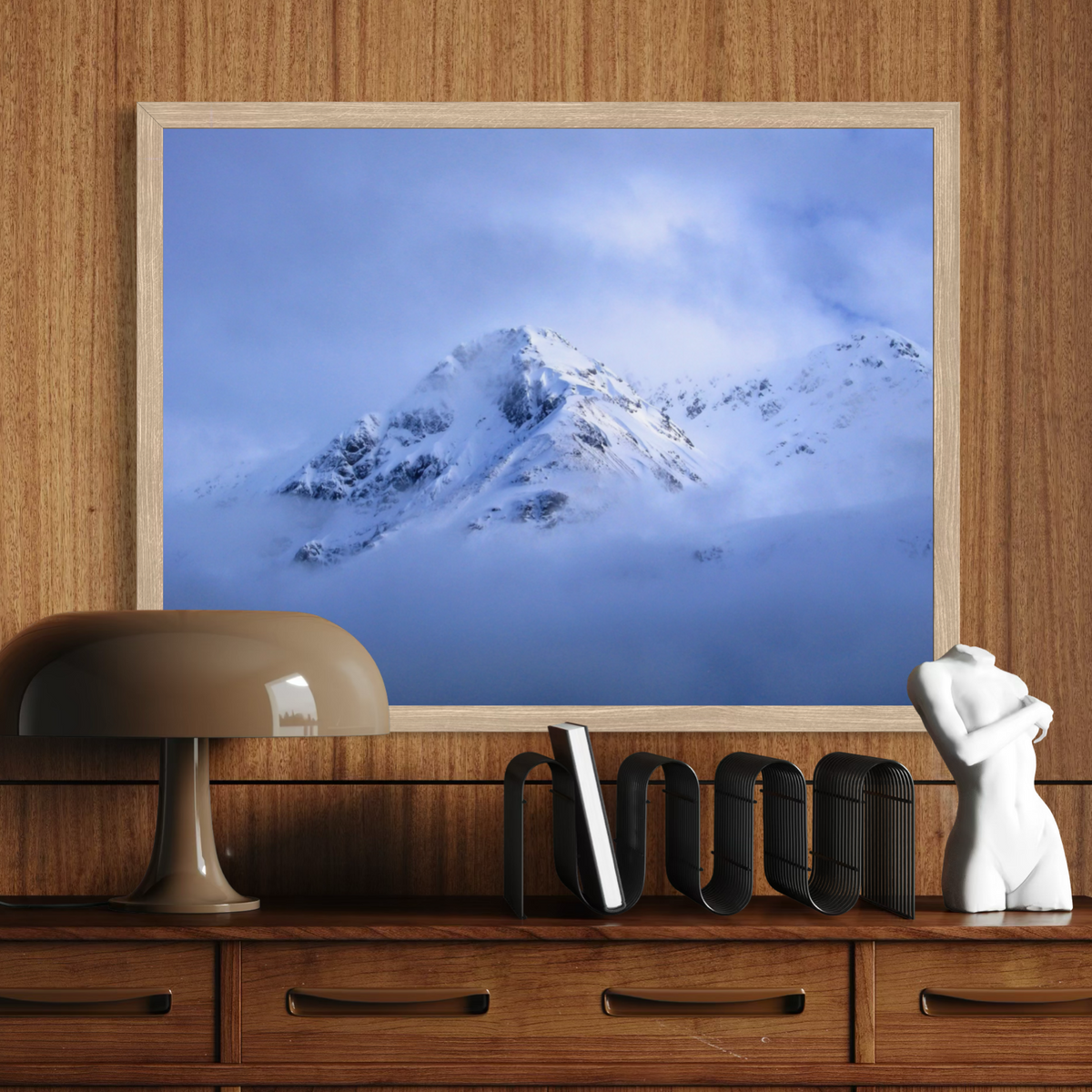 A framed art print depicting a snowy mountain peak under a cloudy blue sky, displayed on a wooden wall above a wooden console table with a lamp, books, and a white figurine.