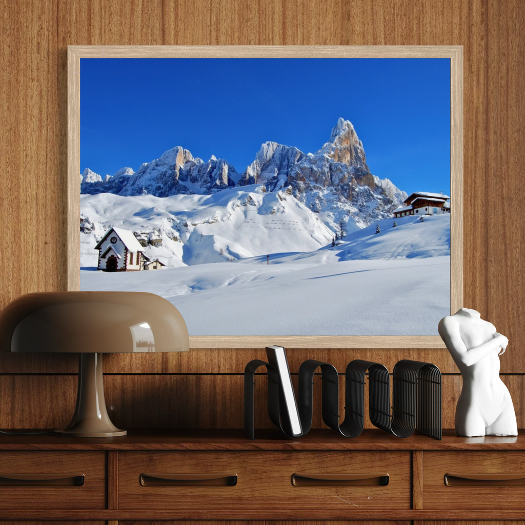 A framed art print depicting a snowy mountain landscape under a clear blue sky, displayed on a wooden wall above a console table with a lamp, bookends, and a small sculpture.