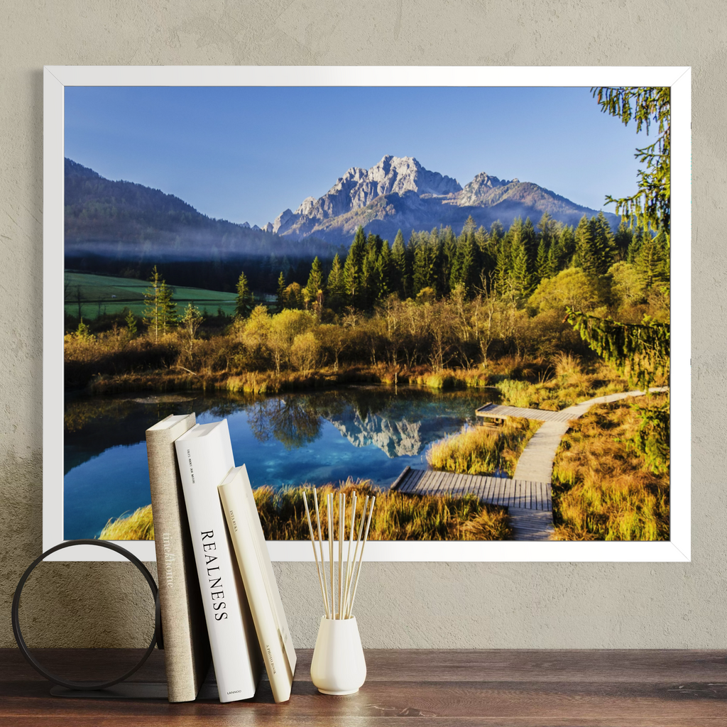 A white framed landscape art print depicting a mountain, forest, and reflective lake, displayed on a wall above a wooden surface with books and a diffuser.