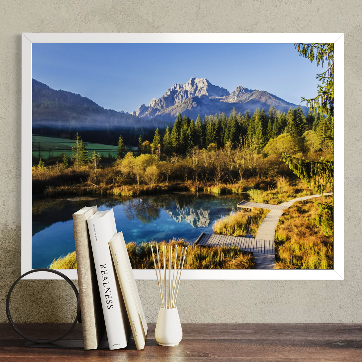 A white framed landscape art print depicting a mountain, forest, and reflective lake, displayed on a wall above a wooden surface with books and a diffuser.