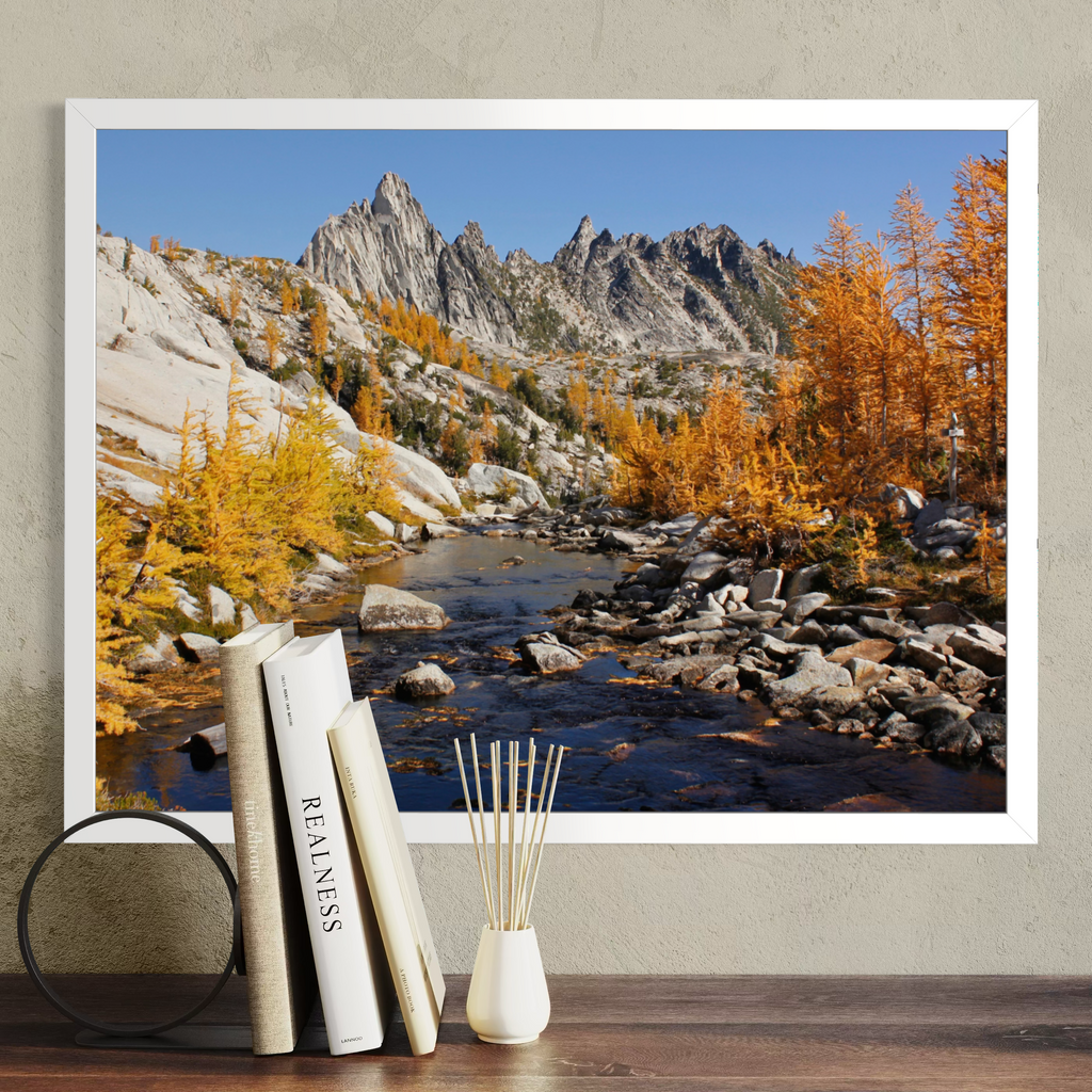 A white framed landscape art print depicting a rocky mountain stream with golden autumn trees, displayed on a wall above a wooden surface with books and a reed diffuser.