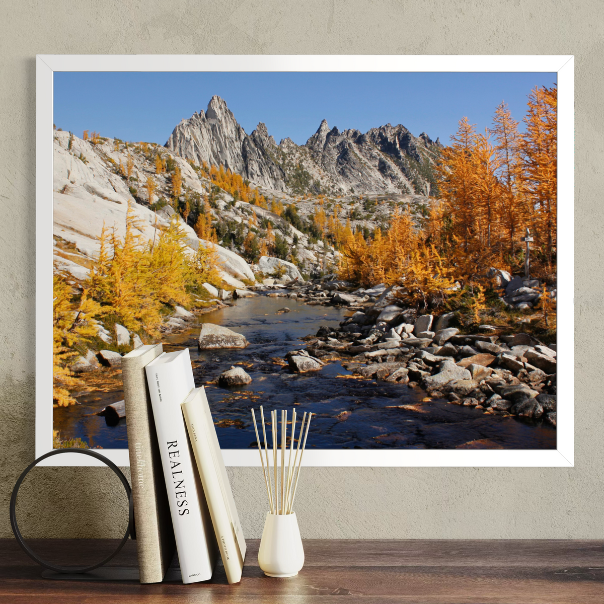A white framed landscape art print depicting a rocky mountain stream with golden autumn trees, displayed on a wall above a wooden surface with books and a reed diffuser.
