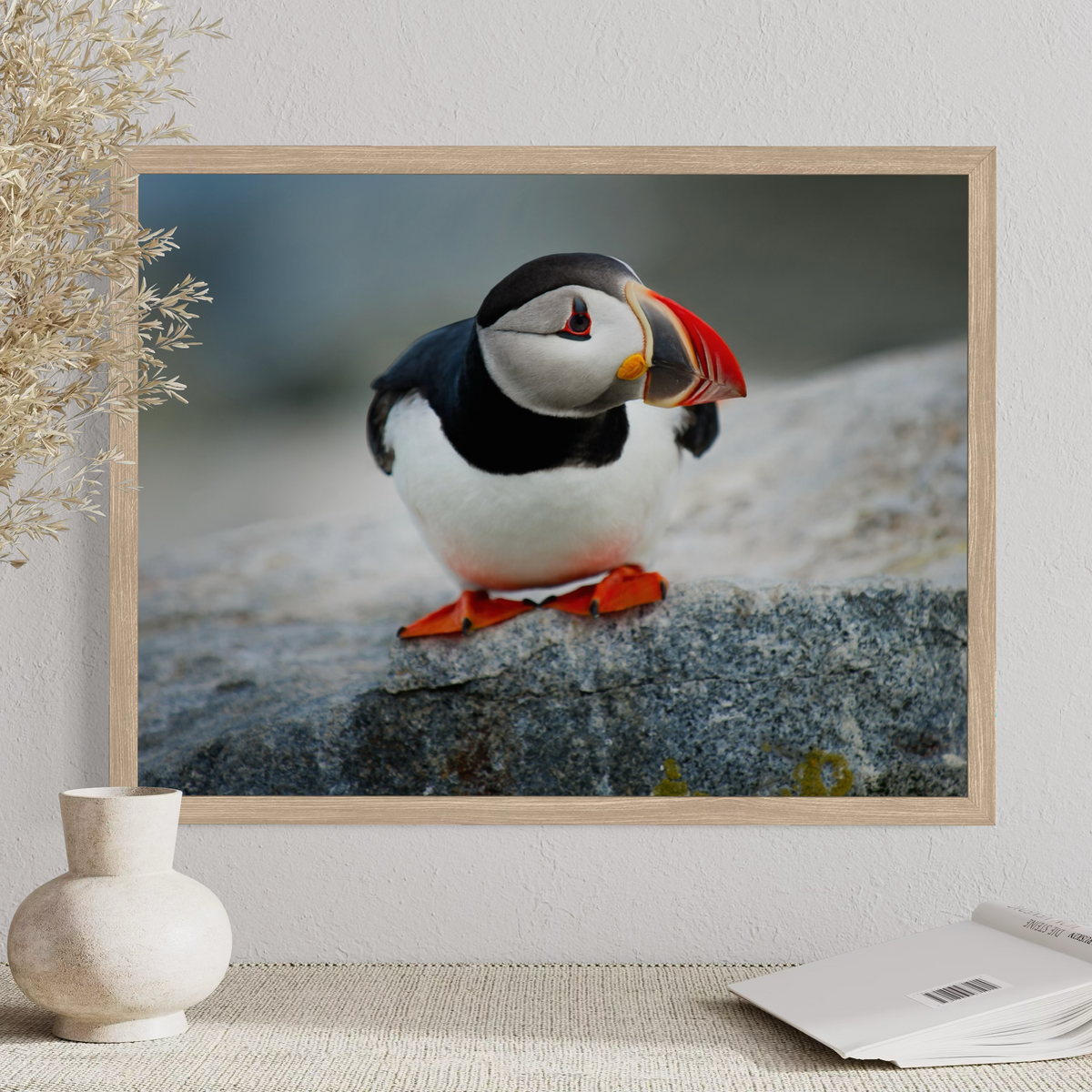 A wooden framed photographic print of a puffin perched on a rock, displayed on a wall above a small vase and an open magazine. The product being sold appears to be the framed wall art itself rather than the bird in real life.