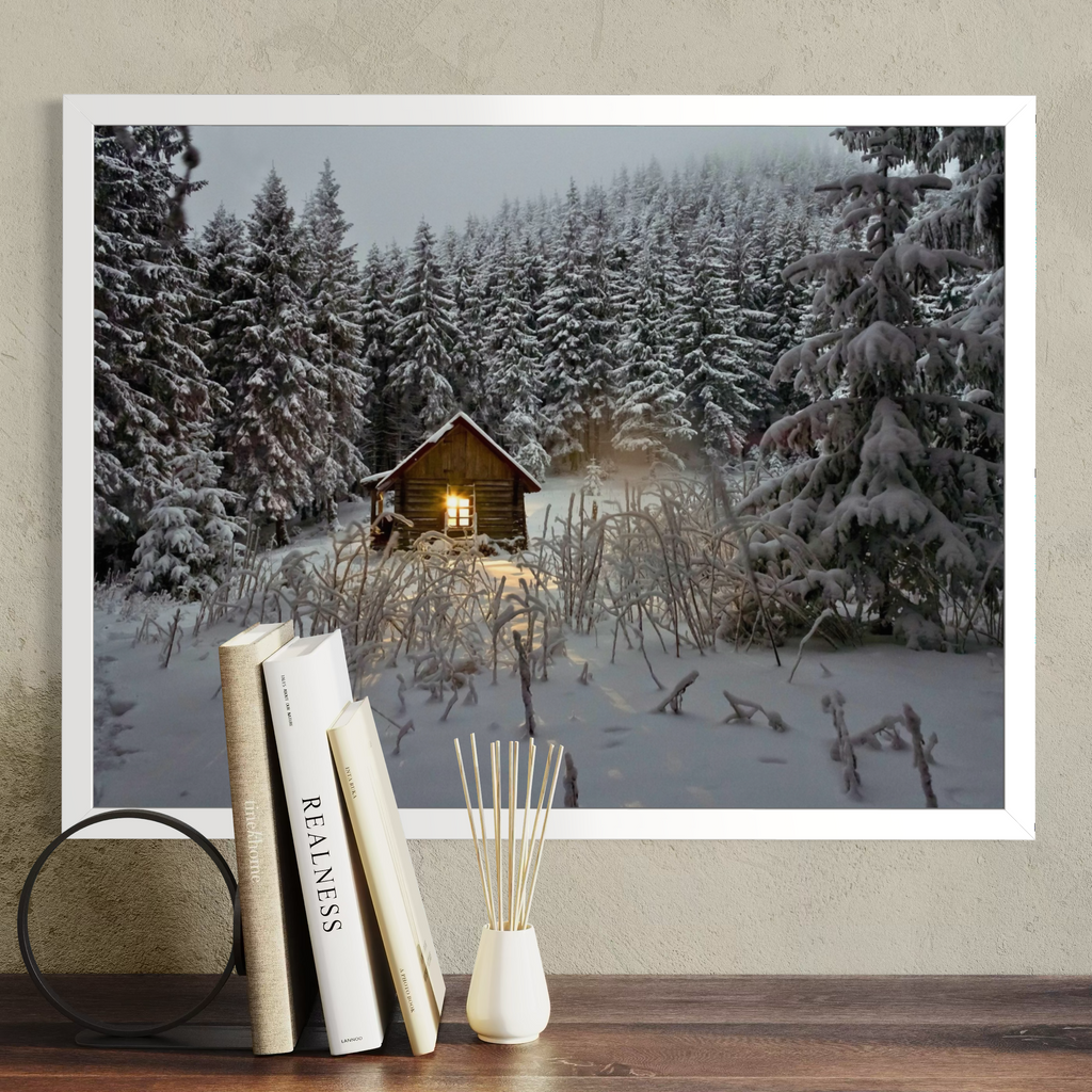 A framed wall art print showing a snowy winter forest with a small cabin and glowing window, displayed above a wooden surface with books and a diffuser against a textured beige wall.